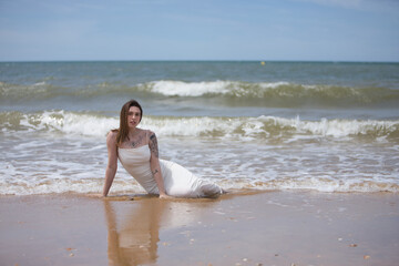 Fototapeta premium Young, beautiful, Hispanic woman, very tattooed, wearing a white dress, sensual and daring, immersed in the water at the edge of the beach, while the wind moves her hair.