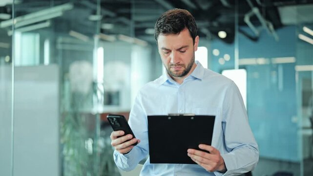 Focused corporate manager or IT specialist looking at mobile screen, verifying project data or documentation. Male employee or freelance worker analyzing report during professional task at workplace