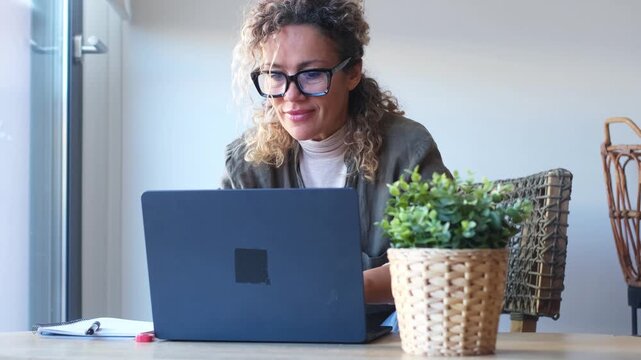 Successful mature businesswoman wearing glasses and smiling while working on a laptop computer, engaged in a video call at her desk in a bright and modern office environment