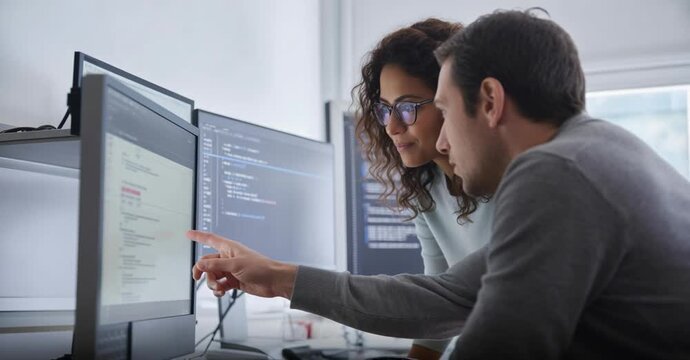 Two diverse professional software developers collaborating and discussing code on a computer screen in a modern tech office. Man points at data, woman observes, showcasing teamwork and innovation in I