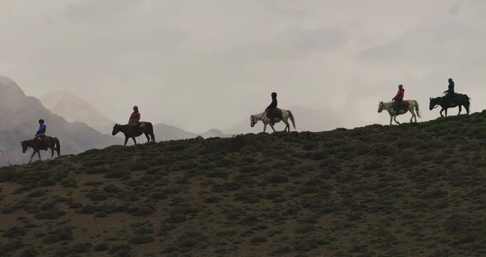 A cinematic, orbiting aerial shot captures a group of horseback riders moving along a mountain slope in Mustang, Nepal, silhouetted against a dramatic backdrop of towering Himalayan peaks. Mustang, Ne