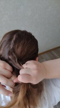 Woman braiding her own long brown wavy hair into a French braid or fishtail plait at home. Close-up over-the-shoulder rear view. Morning beauty routine, hair care, and styling concept video.