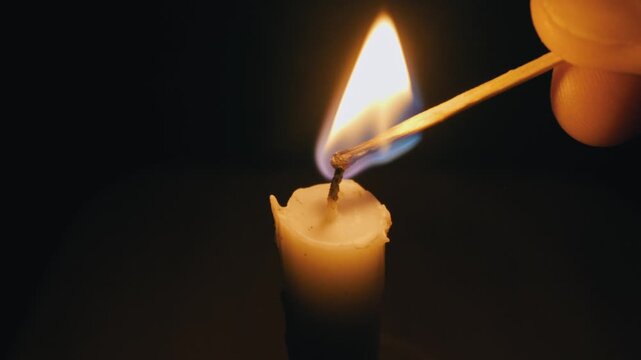 Hand uses a lit wooden match to ignite the wick of a single wax candle. Close-up shot with a completely black background, illuminated only by the warm flame.