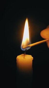 Hand lights a candle wick with a burning match in extreme close-up. Flame illuminates the dark on a black background, creating an intimate, warm glow.