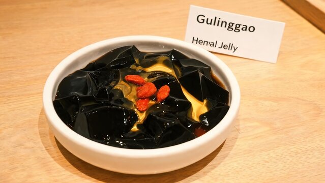 Gulinggao (Guilinggao / Chinese Black Herbal Jelly Dessert) with Honey and Goji Berries in a White Bowl on Wooden Table