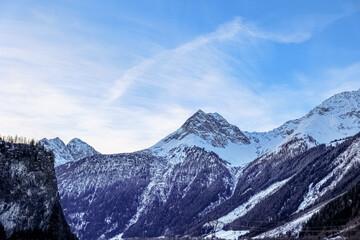 Naklejka premium Snowy mountain peaks and alpine forest landscape under blue sky with wispy clouds, winter nature scenery, the winter nature scenery features rugged rock formations and snow-covered trees
