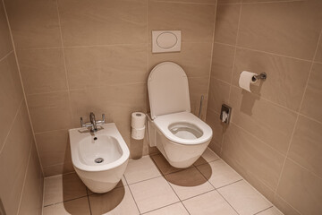 Fototapeta premium Modern bathroom interior with white ceramic toilet and bidet, beige tiled walls and floor, clean restroom design