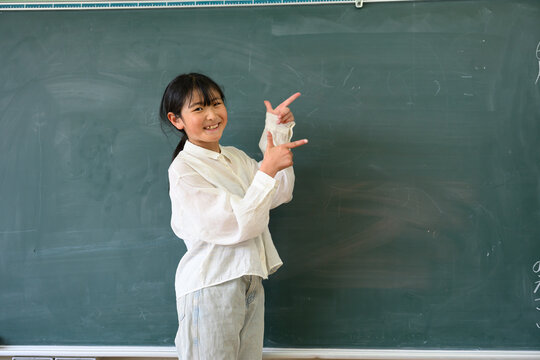 教室の黒板の前で笑顔で指差しをする高学年の女子小学生