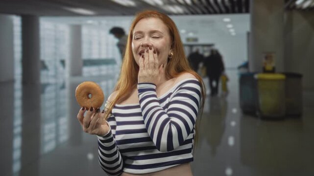 Redhead woman holding a glazed donut, hand covering mouth in a busy airport terminal with travelers and bins visible; playful delight.