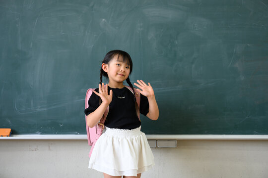 教室の黒板の前でランドセルを背負い両手を広げて笑顔を見せる小学1年生の女の子