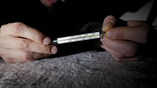 Male hands hold a glass mercury thermometer displaying a high body temperature. Dramatic, high-contrast lighting in a dark room highlights the measurement of the fever.