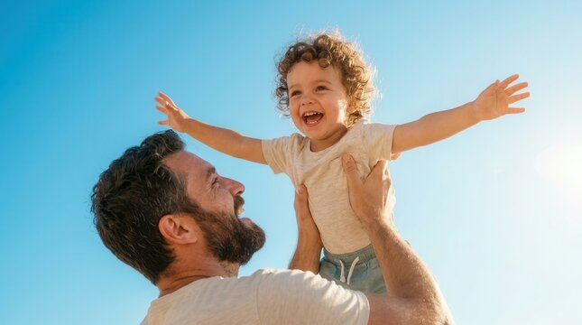 A happy bearded father lifting his curly haired toddler son high against a clear blue sky.