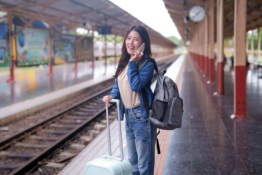 Young Asian woman traveler talking on phone at train station platform with luggage