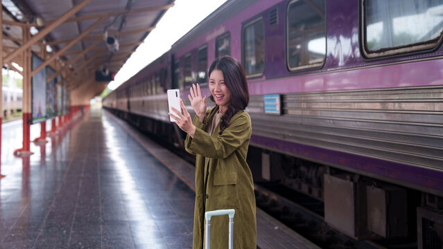 Asian woman traveler waving and video calling on smartphone at train station platform