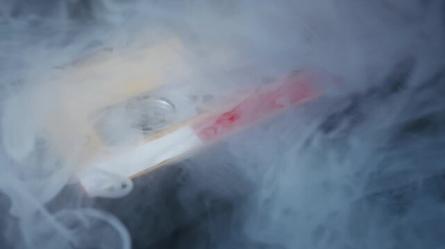 Audio cassette tape with weathered yellow label rests on a dark surface. Mysterious white smoke billows across the frame, obscuring and revealing the object.