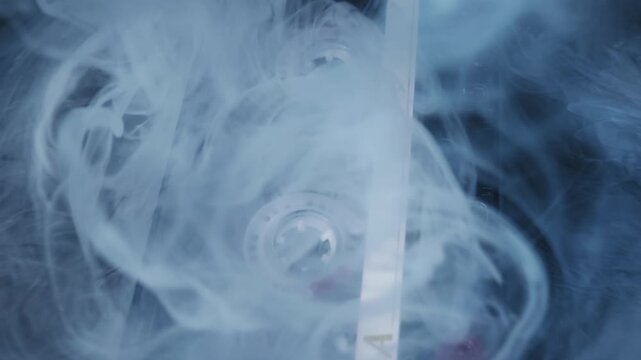 Audio cassette tape with a white label is engulfed by thick smoke on a dark surface. Moody, dramatic lighting and a slow camera pullback create a mysterious and nostalgic atmosphere.