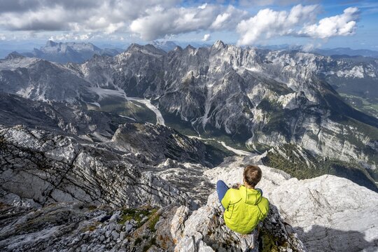 View of Wimbachgries valley and mountain panorama with rocky mountain peak of Hochkalter, mountaineer sitting on a rock at Watzmann Hocheck summit, Berchtesgaden National Park, Berchtesgaden Alps, Bavaria, Germany