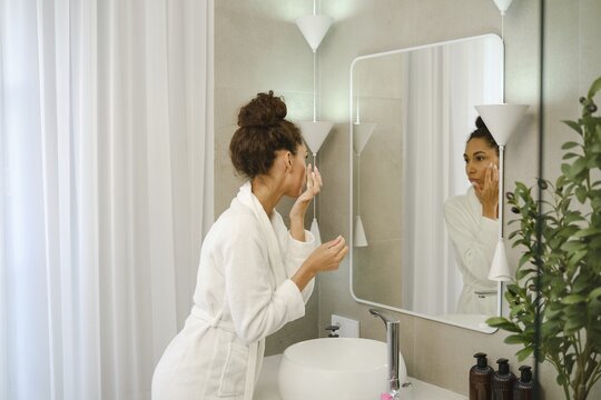 A woman in a white robe is applying skincare in a stylish bathroom. She is focused on her reflection in the mirror, creating a serene and personal moment. Plants add a touch of freshness