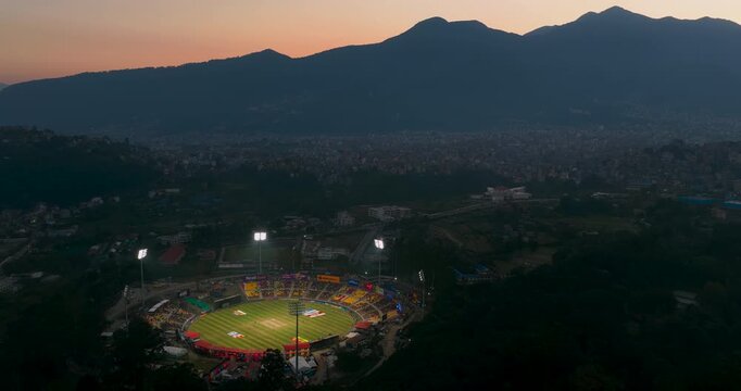 Distant orbiting aerial footage circles high above TIA Ground in Nepal, revealing the cricket stadium illuminated by floodlights against a beautiful hill silhouette and evening sunset.