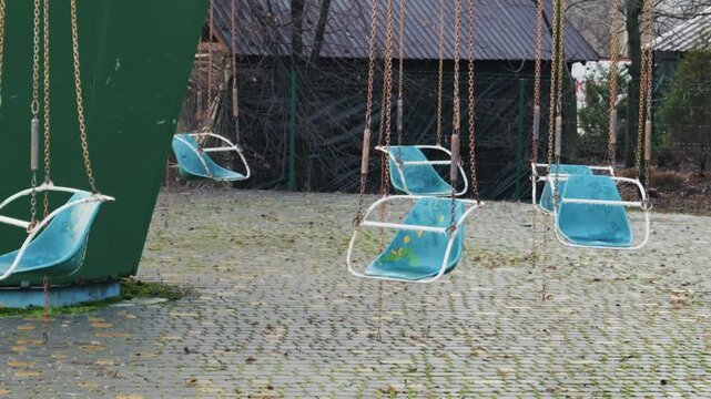 Empty blue seats of a dilapidated chain swing ride hang motionless in a derelict park. A somber and eerie atmosphere is created by the rusty chains and quiet setting.