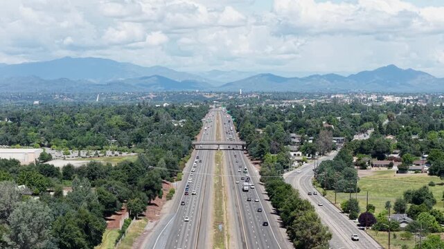 Redding freeway cutting straight through tree-lined suburban sprawl with residential neighborhoods on both sides, Cascade mountain ranges visible on horizon. California, USA. Aerial descend wide shot