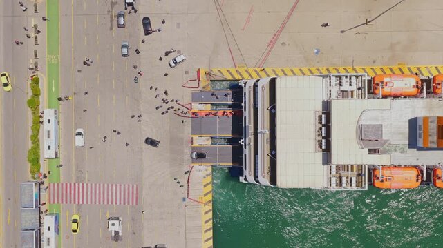 Top-down aerial perspective showing cars and people disembarking from a large passenger ferry docked at a busy harbor, illustrating modern sea transportation and travel logistics