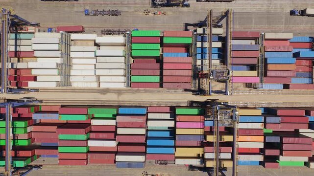 Top-down aerial view of a busy industrial port with large gantry cranes moving along rails, loading and unloading colorful cargo containers stacked in organized rows on the terminal yard