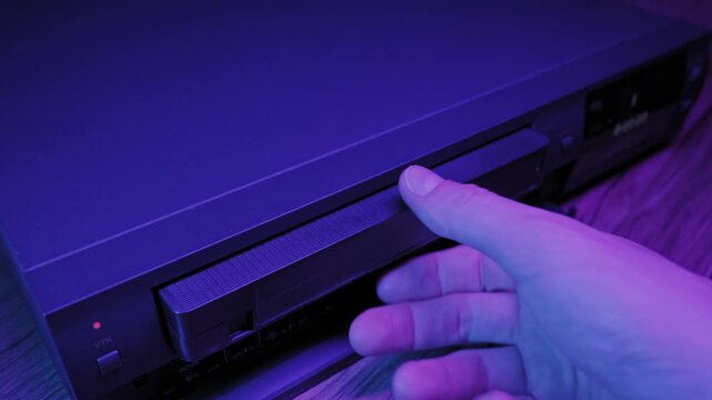 A hand presses the eject button on a vintage VCR player and removes a VHS videocassette tape. The scene is lit with purple neon light. Concept of nostalgia, old technology, and memories.