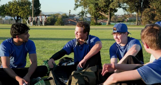 Sitting diverse male teammates forming circle on park sports field, with helmets, water bottles