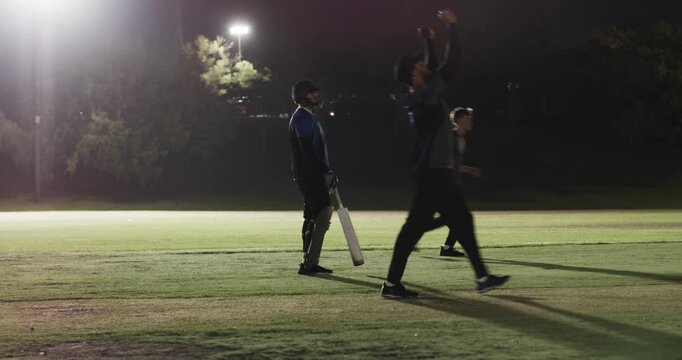 Men playing cricket, bowler bowling appealing wicket, batsman walking off under floodlight
