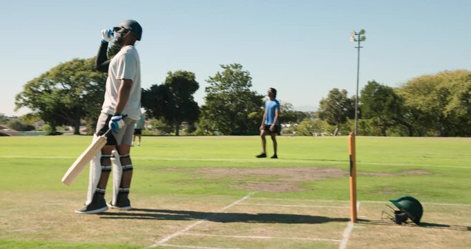 Batsman standing on marked grass pitch holding wooden bat near spare helmet by floodlight tower