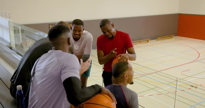 Strategizing diverse male team leaning on glass railing in gym, with basketballs and bag