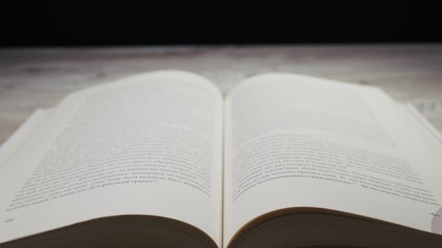 A person's hand flips through the pages of a thick, open book on a wooden table. Concept of studying, knowledge, literature, education, and storytelling.