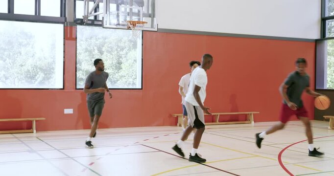 Dribbling basketball under gymnasium hoop, diverse male friends faking move, driving baseline