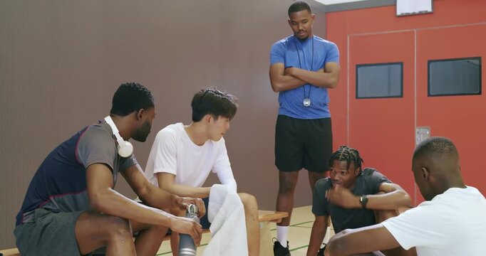 Diverse basketball friends taking water break on bench before coach draping towel