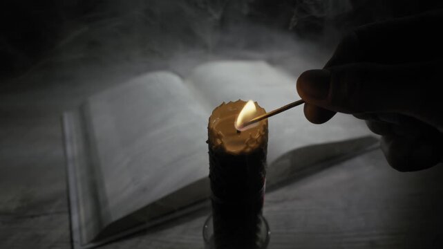 A hand lights a honeycomb beeswax candle with a match next to an open book in a dark, smoky room. Concept of spirituality, seeking knowledge, mystery, and historical atmosphere.