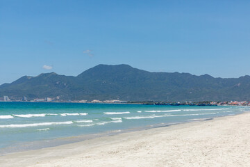 Fototapeta premium Scenic beach view with gentle waves lapping against the shore, mountains rising in the background under a clear blue sky