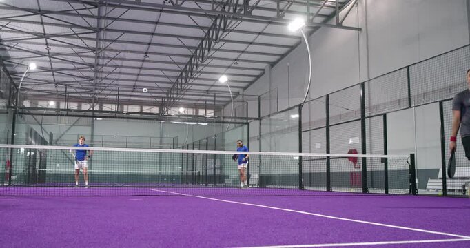 Two male athletes serving underhand shot and exchanging volleys with yellow ball on padel court