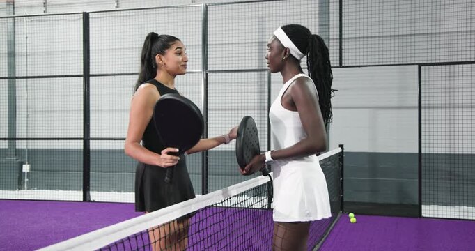 Diverse female athletes tapping paddles approaching padel court net, discussing recent game