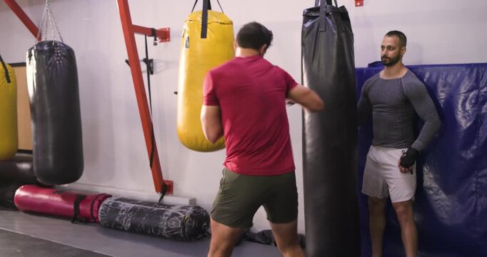 After male coach repositioning bag, trainee executing jabs, roundhouse kicks in gym learning skill