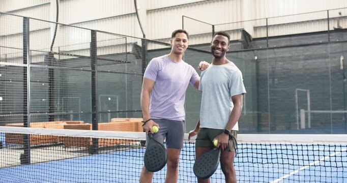Entering padel court with rackets, balls, diverse male friends smiling, fist bumping, chatting