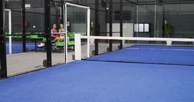 Female player on padel court awaiting shot, swinging backhand, female duo cheering, copy space