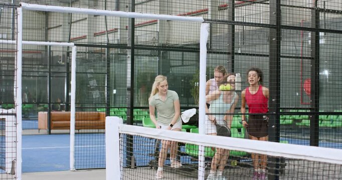 Women friends chatting lifting balls from ball hopper grabbing rackets entering padel court