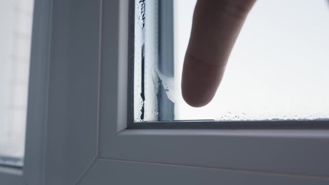 A male finger touches water condensation on a white window frame, revealing heavy moisture buildup. Concept of household humidity problems and poor insulation in winter.