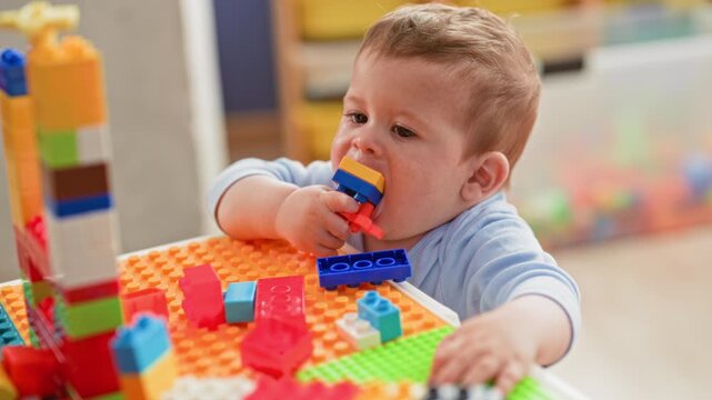 Boy chewing a colorful plastic building block with his hand at a playroom table, reaching for stacked blocks; childhood curiosity.