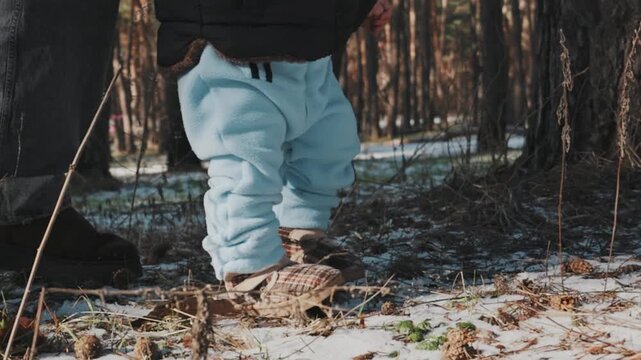 Close-up of a baby's legs in blue pants taking unsteady first steps on snowy ground with parental support. Concept of child development, family support, and new beginnings in nature.