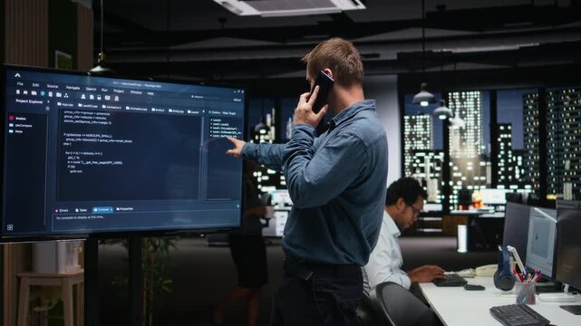 Manager analyzes code on screen, makes phone call to ask for better automation of business tasks in workspace. Supervisor talking on smartphone checks programming language scripts on monitor.