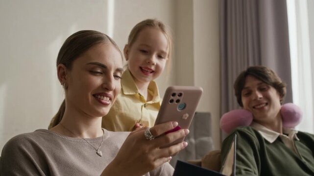 Medium shot of Caucasian young adult woman checking smartphone and passport while child and father relaxing in background. Ideal for family travel apps and hotel booking services