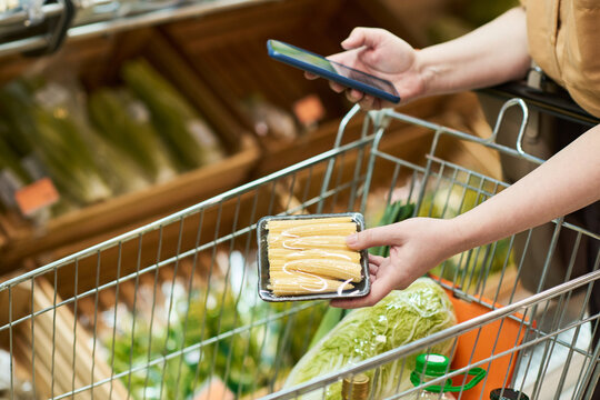 young woman shopping for groceries, scanning packaged corn with smartphone beside cart in supermarket produce aisle. Useful for retail technology, smart shopping, consumer app marketing