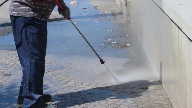 Anonymous city contractor in uniform performing surface restoration by deep cleaning stone masonry parapet wall and urban walkway with heavy duty power washer under bright summer sunlight mist